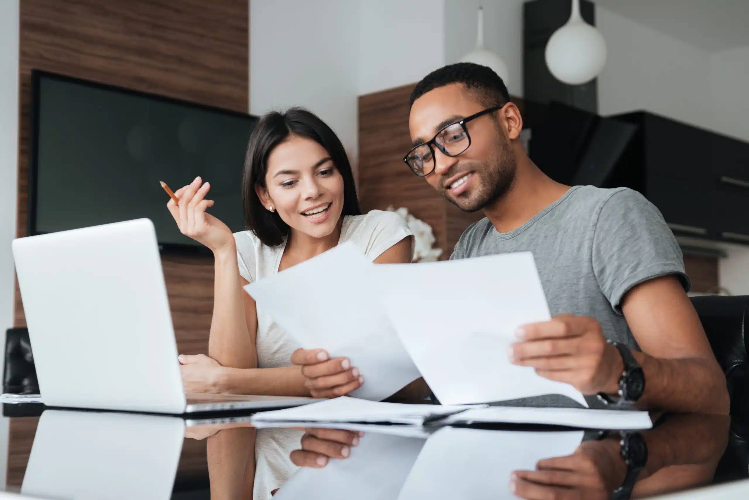 Photo of cheerful loving young couple using laptop and analyzing their finances with documents. Look at papers.