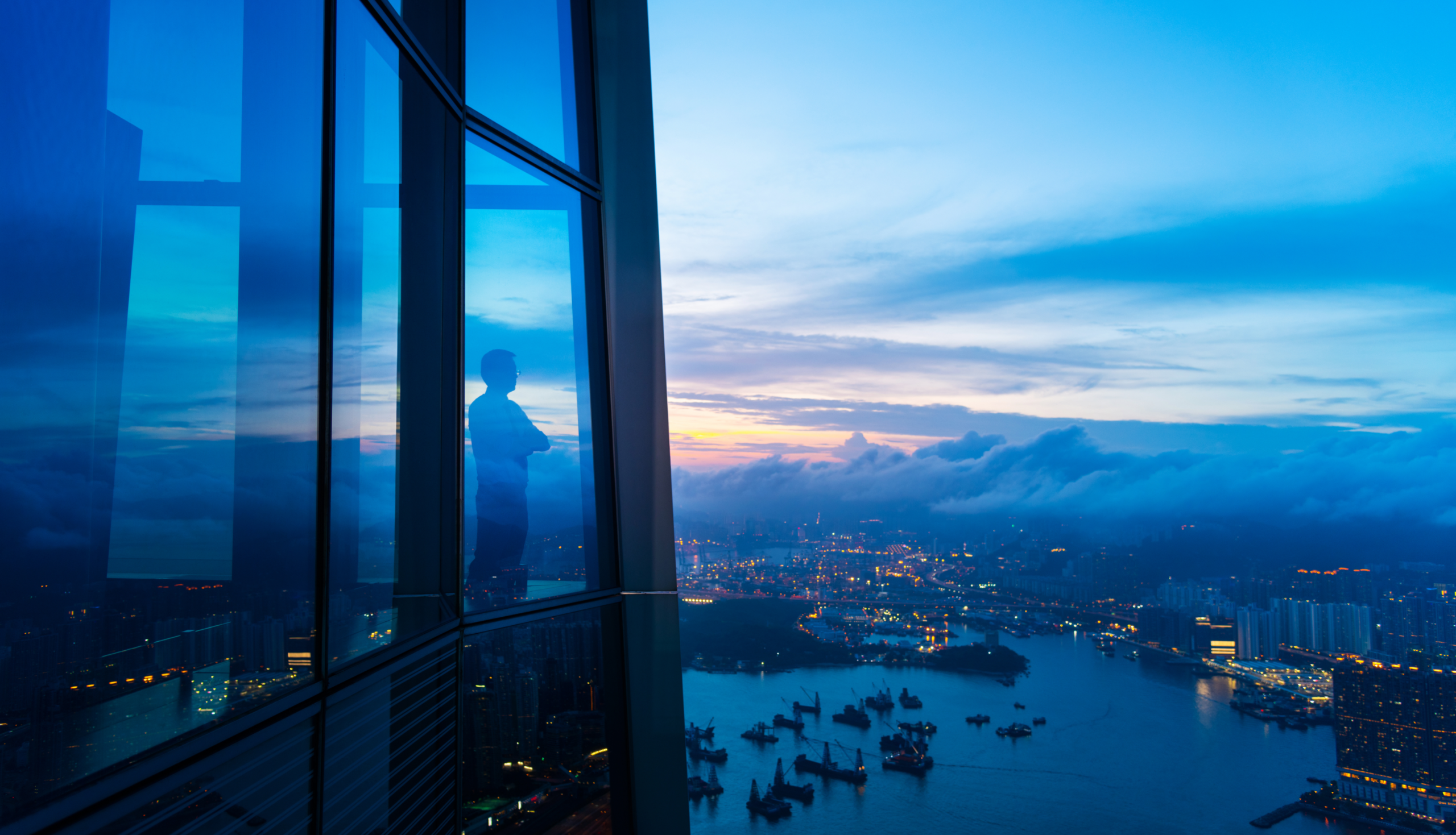 A foreign-born wealthy professional in a suit looking out of a high-rise office building onto a waterfront, symbolizing global mobility and cross-border opportunity