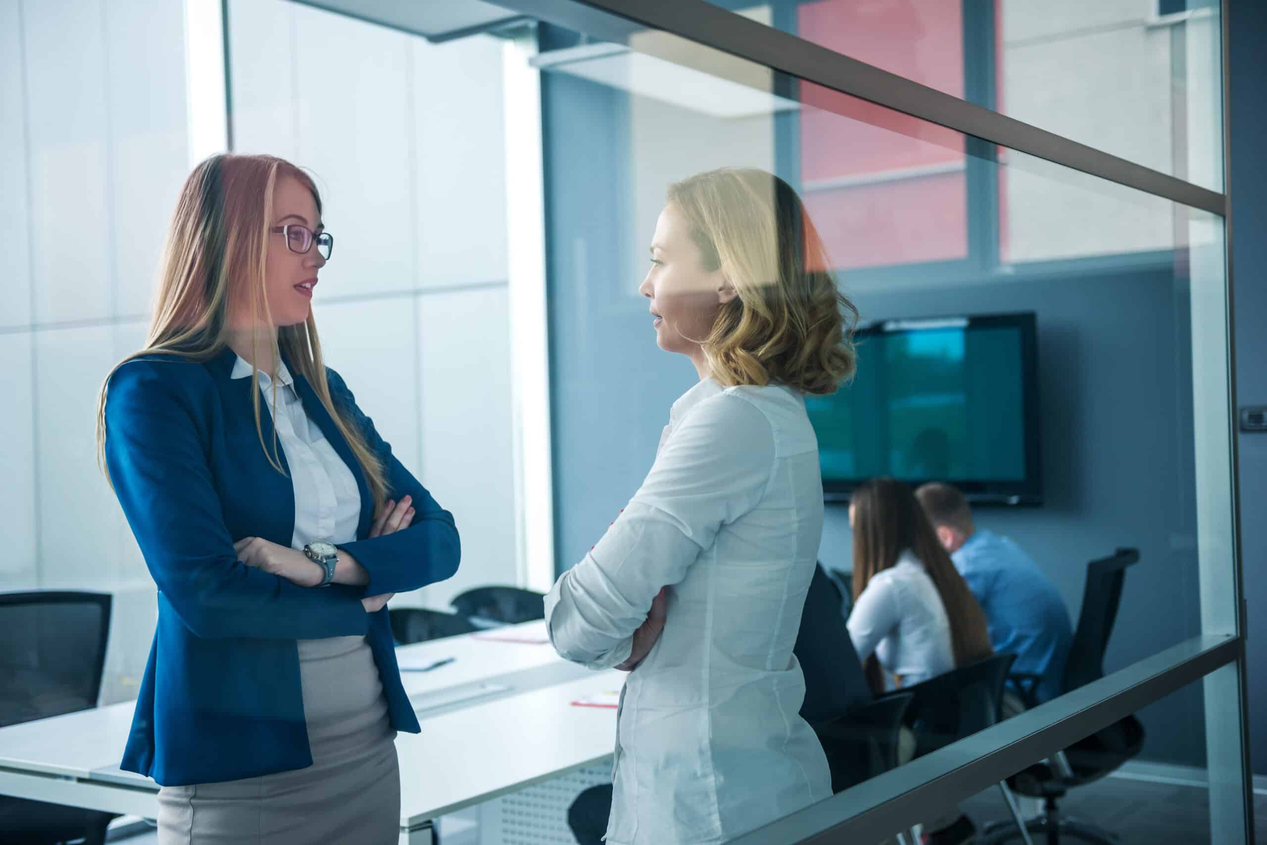Two women standing together in a modern office discussing wealth management strategy and relationship intelligence for UHNW client acquisition.