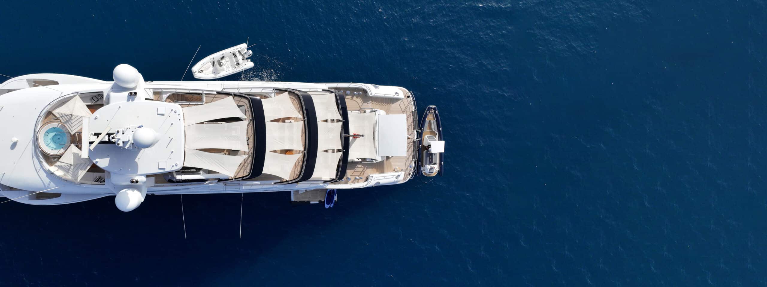 Aerial view of a white luxury yacht cruising on deep blue water, with a small tender alongside.