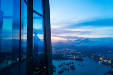 A foreign-born wealthy professional in a suit looking out of a high-rise office building onto a waterfront, symbolizing global mobility and cross-border opportunity